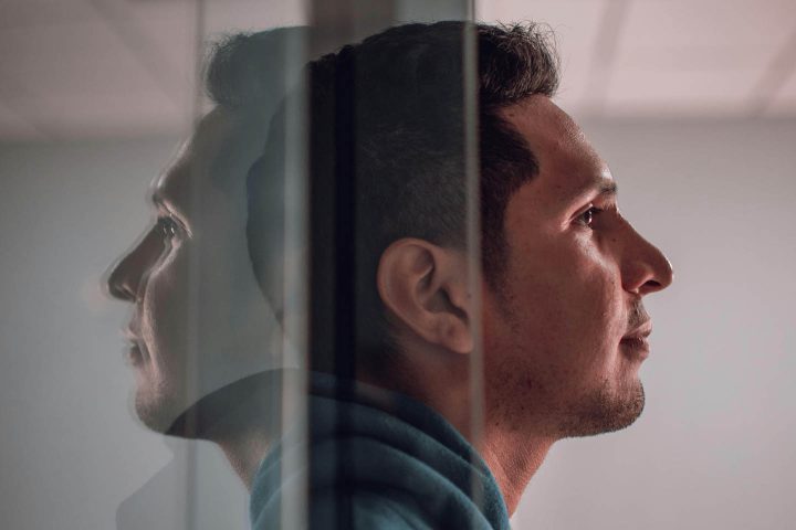 Een man die door een glazen deur naar buiten stapt, terwijl de weerspiegeling van zijn gezicht achterblijft.
